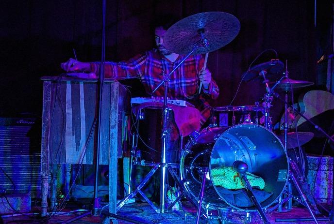 Person playing drums onstage with purple and green lights around them.