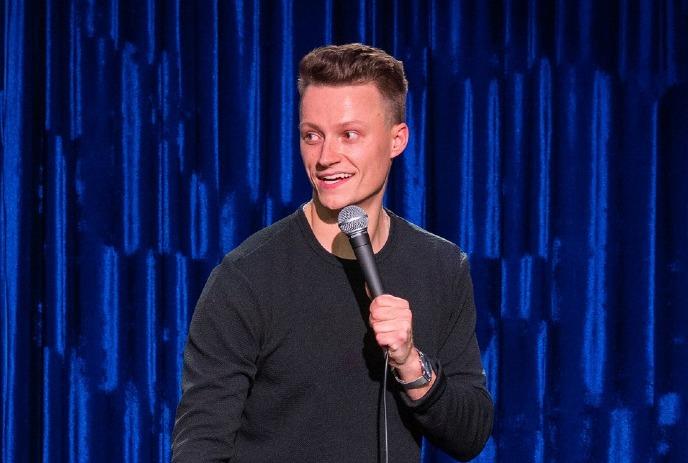 Andrew Packer performing stand up comedy in front of a blue curtain.