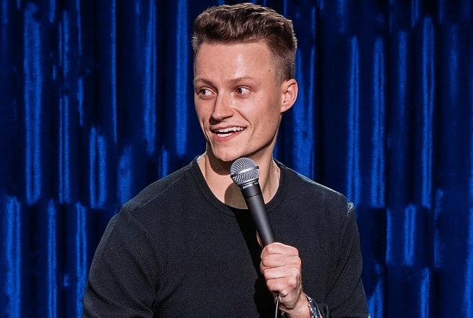 Andrew Packer performing stand up comedy in front of a blue curtain.