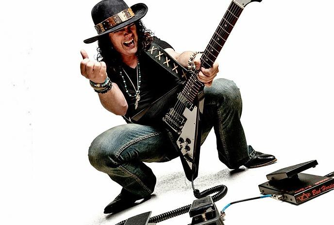 Anthony Gomes  in a dynamic pose playing an electric guitar, surrounded by pedals, and wearing a hat.