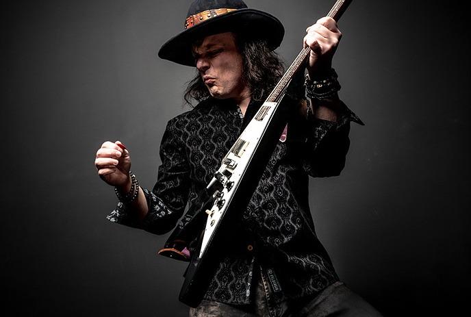 Anthony Gomes playing an electric guitar, wearing a black patterned shirt, dark pants, and a wide-brimmed hat.