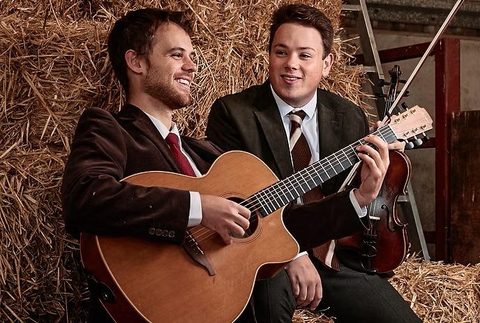 Two suited individuals seated on hay bales in rustic barn one playing guitar beside resting violin on straw.