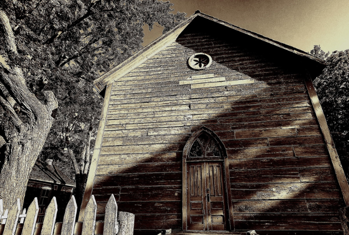 Sepia-toned photograph of an old wooden building with a pointed roof and a gothic-style arched doorway.