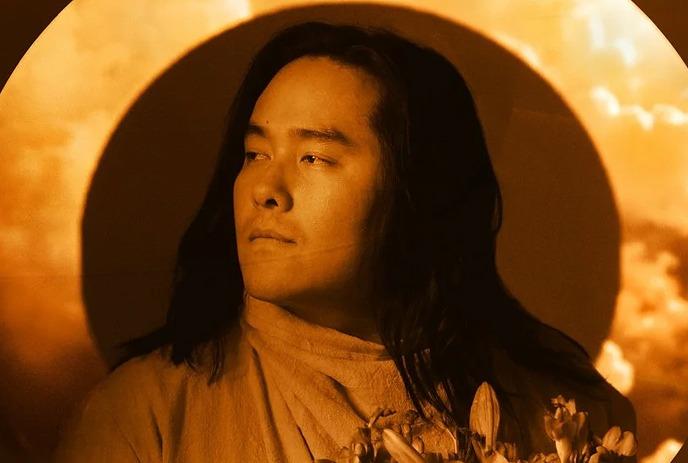 Aysanabee with long hair holds flowers, standing against a glowing orange circle backdrop.