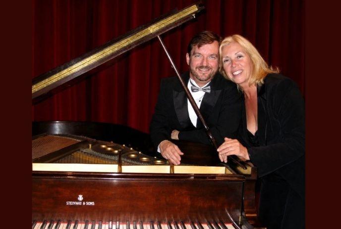 Clark Bryan & Marion Miller poses beside a grand piano in formal attire, with elegant red curtains on the background.