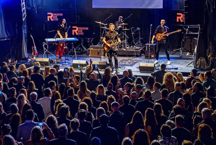 Crowd faces bright stage with lights and fog as people raise hands in energetic live music scene.