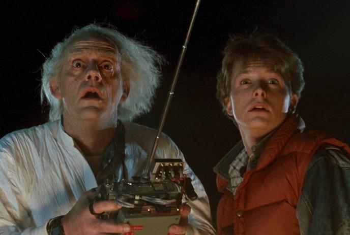 A scene from Back to The Future where two individuals are looking forward with one with a controller under dark background.