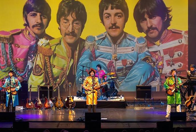 A tribute band performs on stage in colorful Sgt. Pepper uniforms, set against a large backdrop of The Beatles.