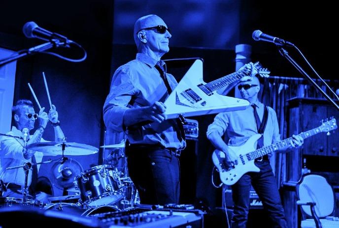 A band performs on stage with drummer, guitarist, and bassist—white instruments gleam under spotlights.