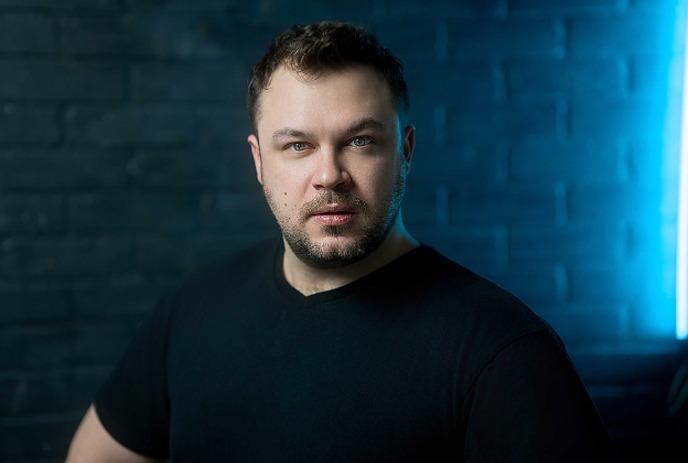 Comedian Ben Bankas stands against a brick wall with blue lighting, wearing a black shirt.