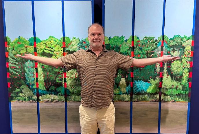 Ben Benedict standing with arms outstretched before tall vivid forest panels with bright greens, blues and reds.