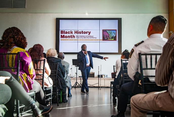 Man in suit speaking at Black History Month event, gesturing to screen in front of seated audience.