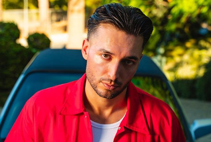 BIJOU wearing a white tank top and glasses, posing for the camera in front of a blue background.