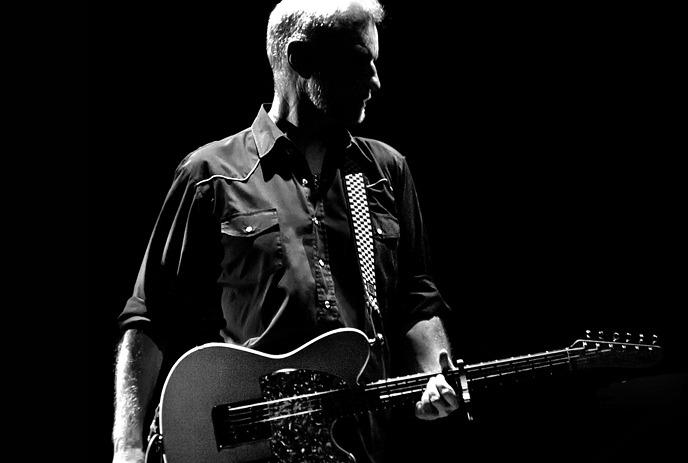 A black and white photo of Billy Bragg, standing in the spotlight holding a guitar.
