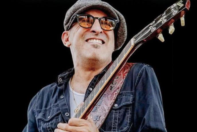 Guitarist in cap and sunglasses plays onstage with a faded larger image of him in the background.