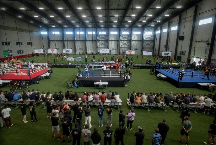 Indoor sports venue with multiple boxing rings where athletes compete as spectators watch from benches nearby.
