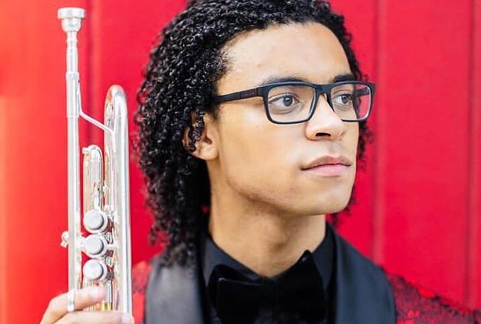 Will Leathers holding a trumpet, dressed up wearing glasses, in front of a red background.