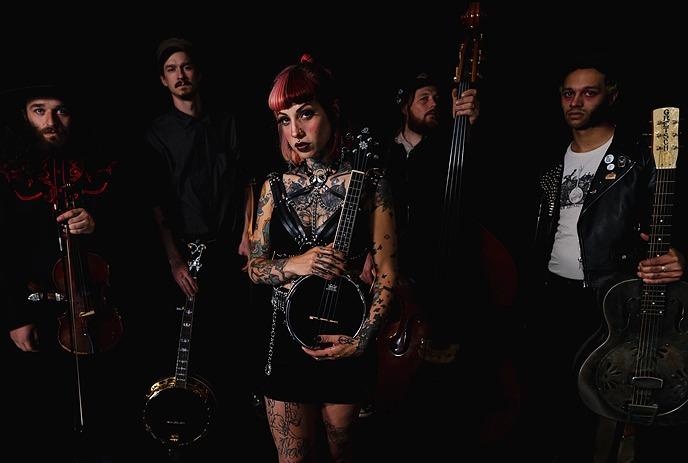 The Bridge City Sinners pose with a violin, banjo, upright bass, and guitar against a dark background.