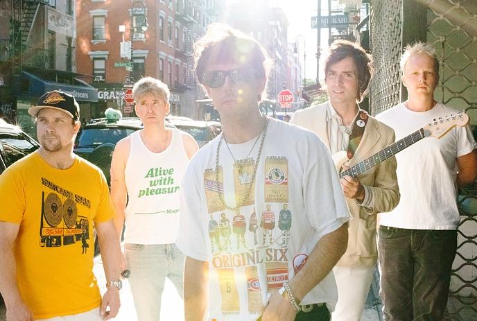 A group photo of the band The Brokes standing on a city street with one member holding a guitar.