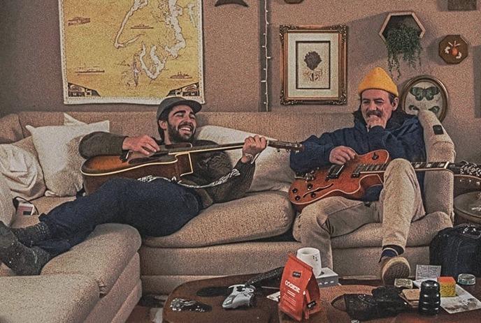 An illustration of two skeletons sitting on a couch, playing guitars in front of mountain landscape wallpaper.