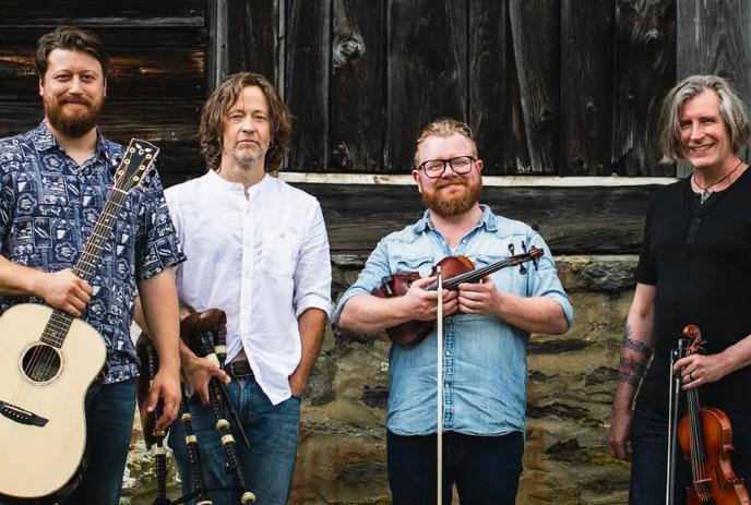 Four musicians standing before rustic wooden stone wall each holding instrument in natural outdoor setting.
