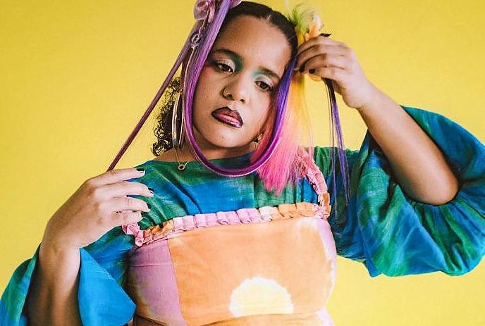 Person with colourful hair and a bright green and turquoise top in front of a yellow background.