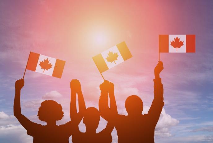 Silhouettes of three people joyfully raise Canadian flags against a vibrant sunset sky with scattered clouds.