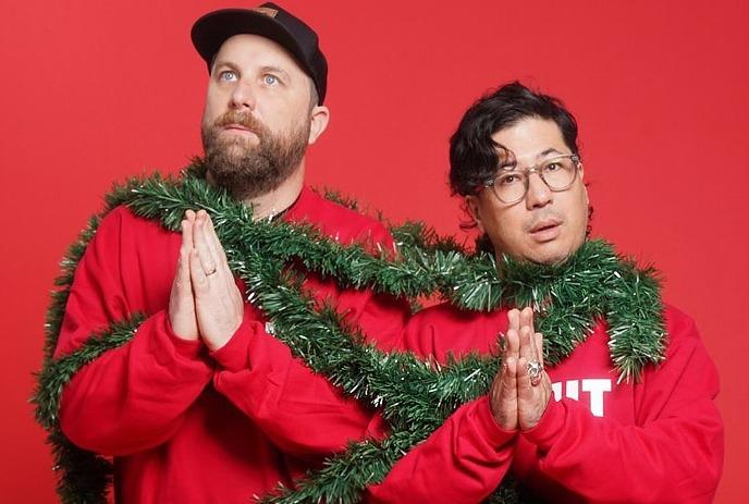 Two artists in festive duo in red sweaters and green garland beam joyfully before a red plaid crowd-filled celebration scene.