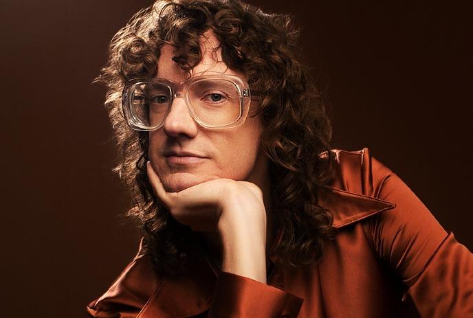Chris Fleming, with curly dark hair and glasses, wears a white shirt, gazing thoughtfully off-camera against teal background.