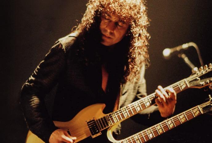 Musician with long curls plays sunburst electric guitar on dim stage lit by green light.