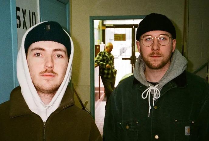 Two young men of Cleopatrick stand on a dimly lit street at night. Both wear casual hoodies, with one in a denim jacket.