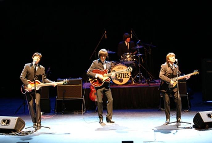 Four musicians perform in suits on stage, playing guitars and drums in front of a