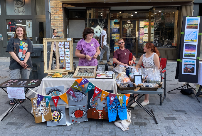 Artists display colorful prints and crafts at an outdoor booth while people browse the vibrant setup.