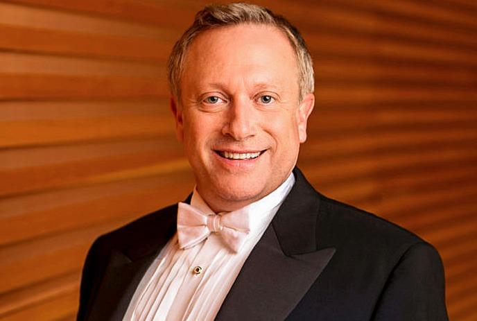 A smiling artist in a tuxedo stands before a blue backdrop decorated with musical notes.