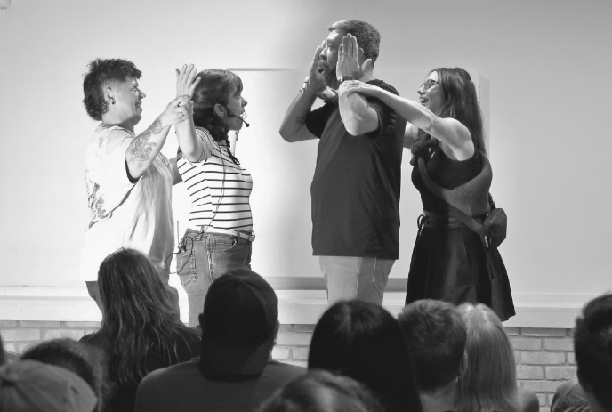 Live performance with four people on stage, palms touching, audience seated and watching in black-and-white frame.
