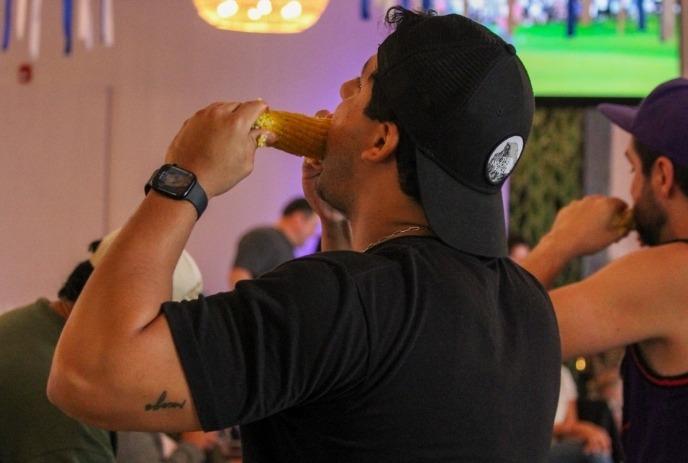 A person in a black cap and shirt eats corn on the cob in a lively indoor setting with others.