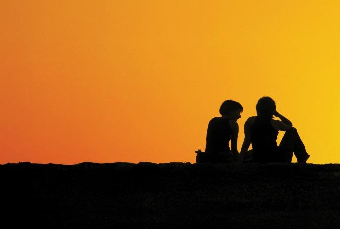 Silhouetted figures of two people sitting on a ledge, gazing at a vibrant orange sunset.
