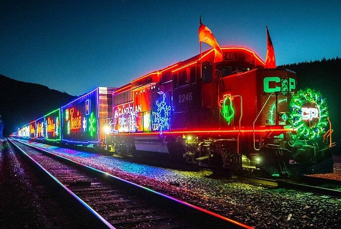 Holiday train glows with festive lights, peace sign, Santa, and trees; snow-covered track at night.