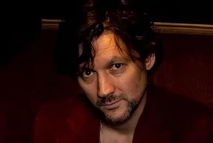 Craig Jamieson sits in dim light, wearing a red jacket and white tee, looking straight ahead.