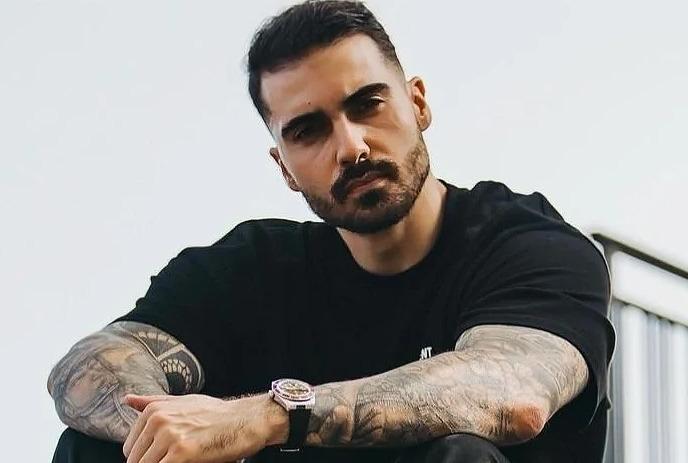 Crusy sitting down on steps with his arms crossed looking at the camera, with a white background.