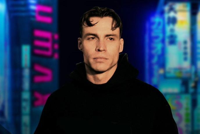 Artist in black hood before vibrant neon city signs glowing at night in a colorful urban street scene.