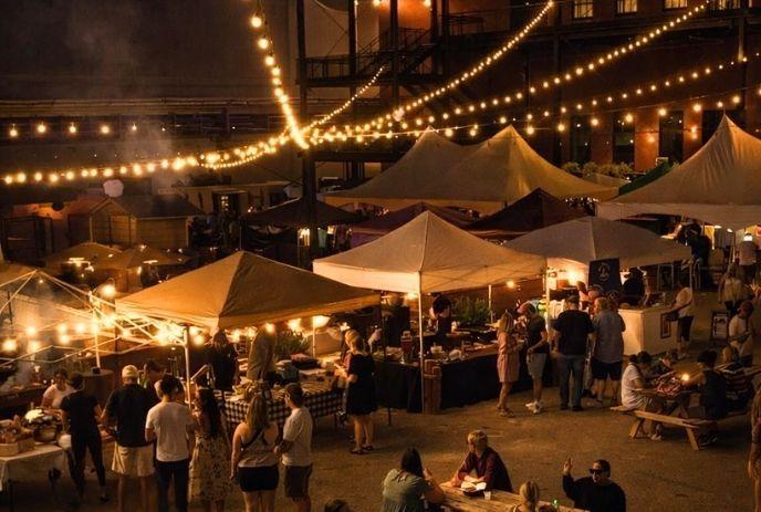 Night market with warm string lights, tents, and people eating, walking, and gathering in a lively courtyard.