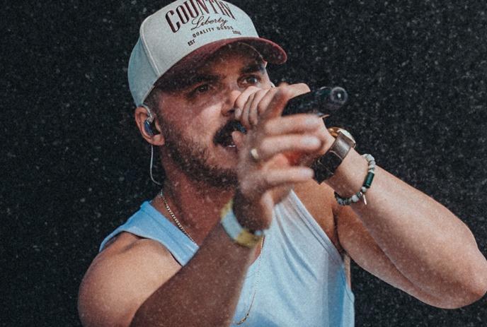 Performer in a white tank top and cap sings into a microphone while pointing toward the audience.