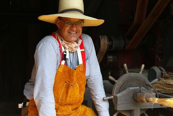 A person in a straw hat and leather apron stands in a barn by a threshing machine with straw.