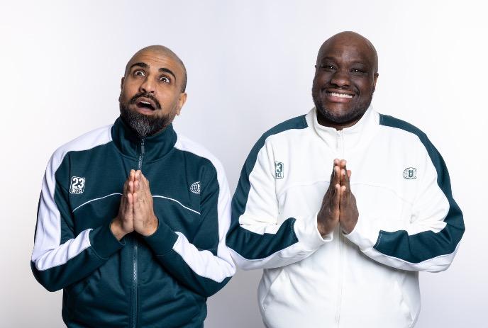 Two comedians artists in matching sporty jackets stand side by side with hands pressed together in a calm gesture.