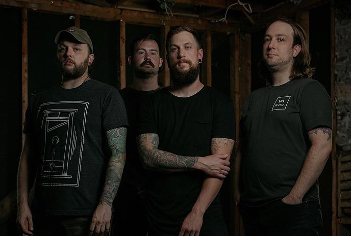 Four men of the band Dead Days stand together in dim lighting, gazing up.