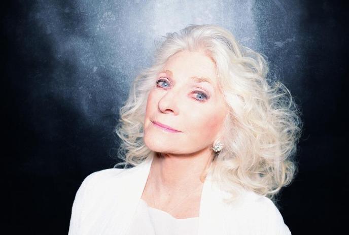 Judy Collins with expressive blue eyes and styled white hair, wearing a white outfit.