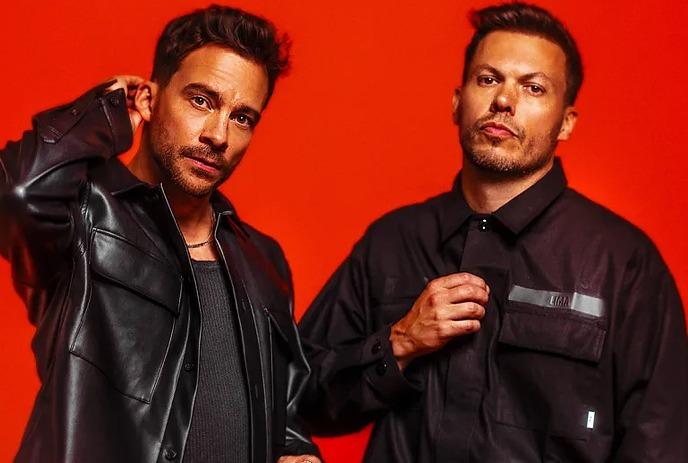 The two band members of Delta Heavy posing in front of the camera wearing black coats, in front of a red background.