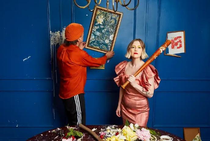 Person in orange hangs a floral painting on a blue wall while another in pink holds a bat, with a messy flower-strewn table below.