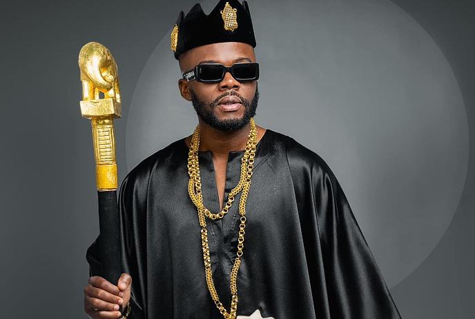 DJ Kerozen wearing sunglasses and holding a staff in front of a hazy pink background.